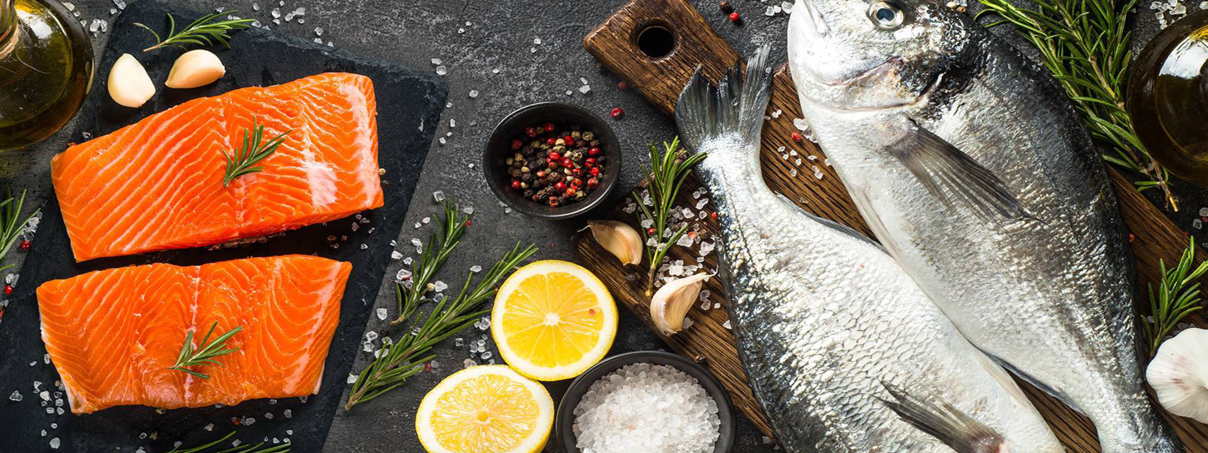 Filetti di salmone e pesce intero fresco con limone, rosmarino, aglio, sale e pepe su sfondo scuro.