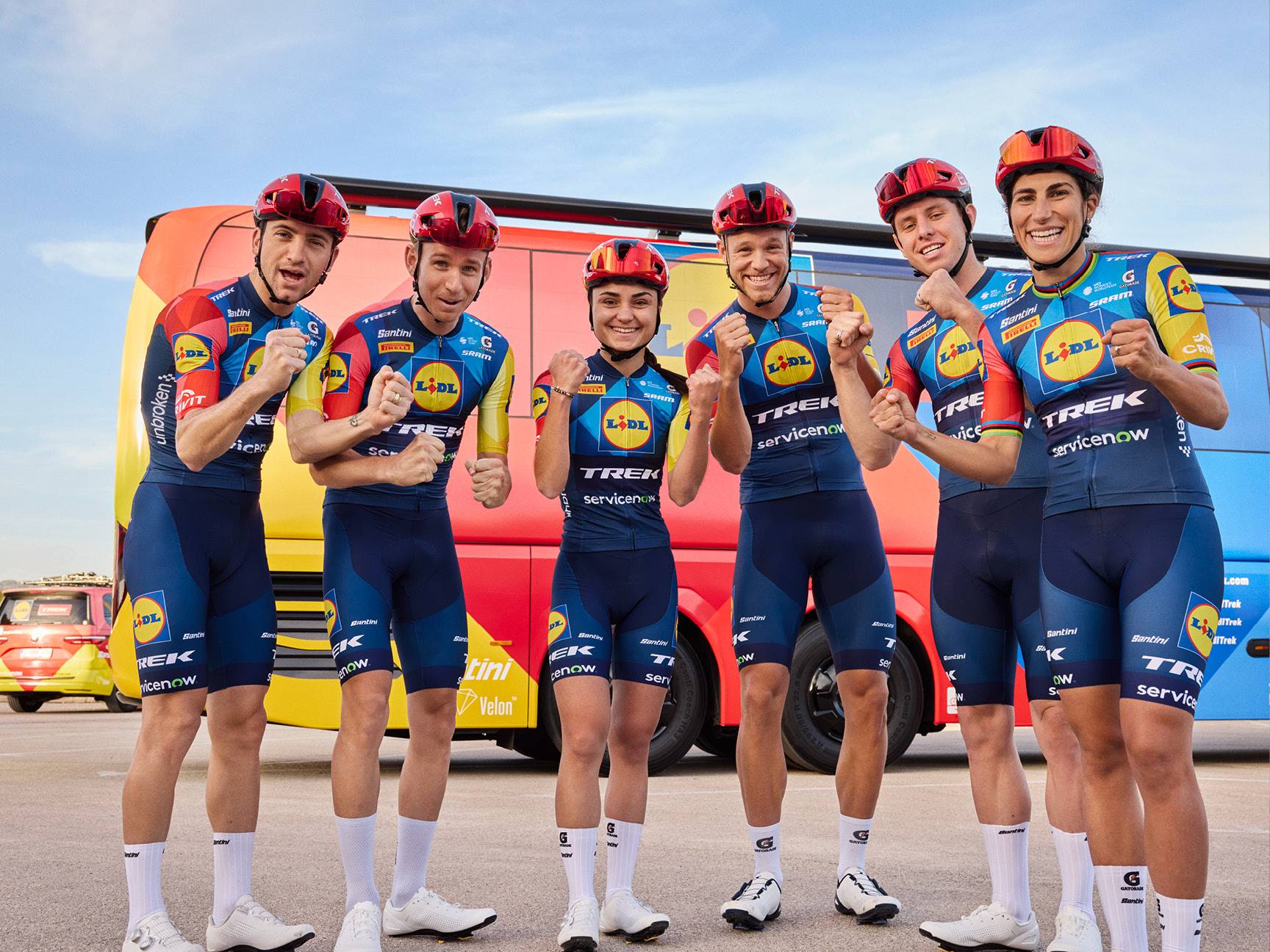 Sei ciclisti in abbigliamento sportivo blu e giallo con caschi rossi, in posa davanti a un autobus.