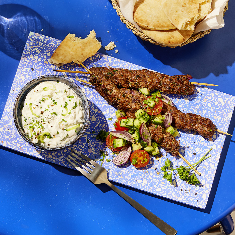 Spiedini di carne macinata con salsa tzatziki, insalata di pomodori e cetrioli, e pane pita.