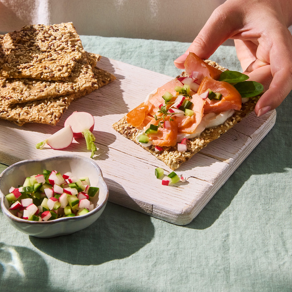 Cracker di semi con salmone affumicato, formaggio cremoso e verdure tritate, tenuto da una mano.