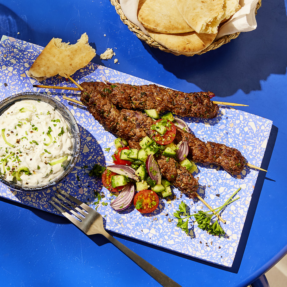 Spiedini di carne macinata con salsa tzatziki, insalata di pomodori e cetrioli, e pane pita su un tavolo blu.