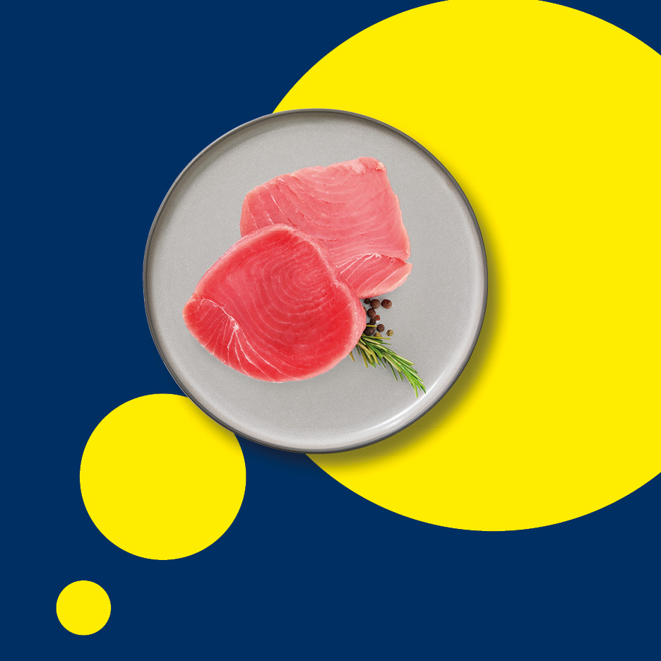 Due filetti di tonno crudo su un piatto grigio, con rosmarino e pepe, su sfondo blu e giallo.