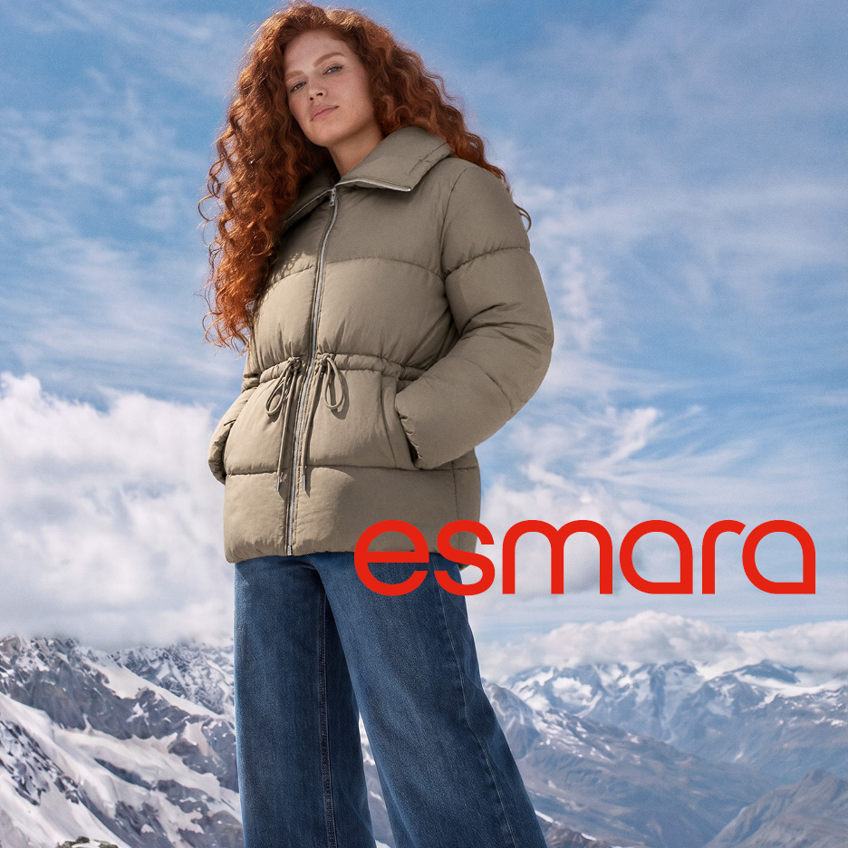 Donna con giacca imbottita beige e jeans su uno sfondo di montagne innevate.