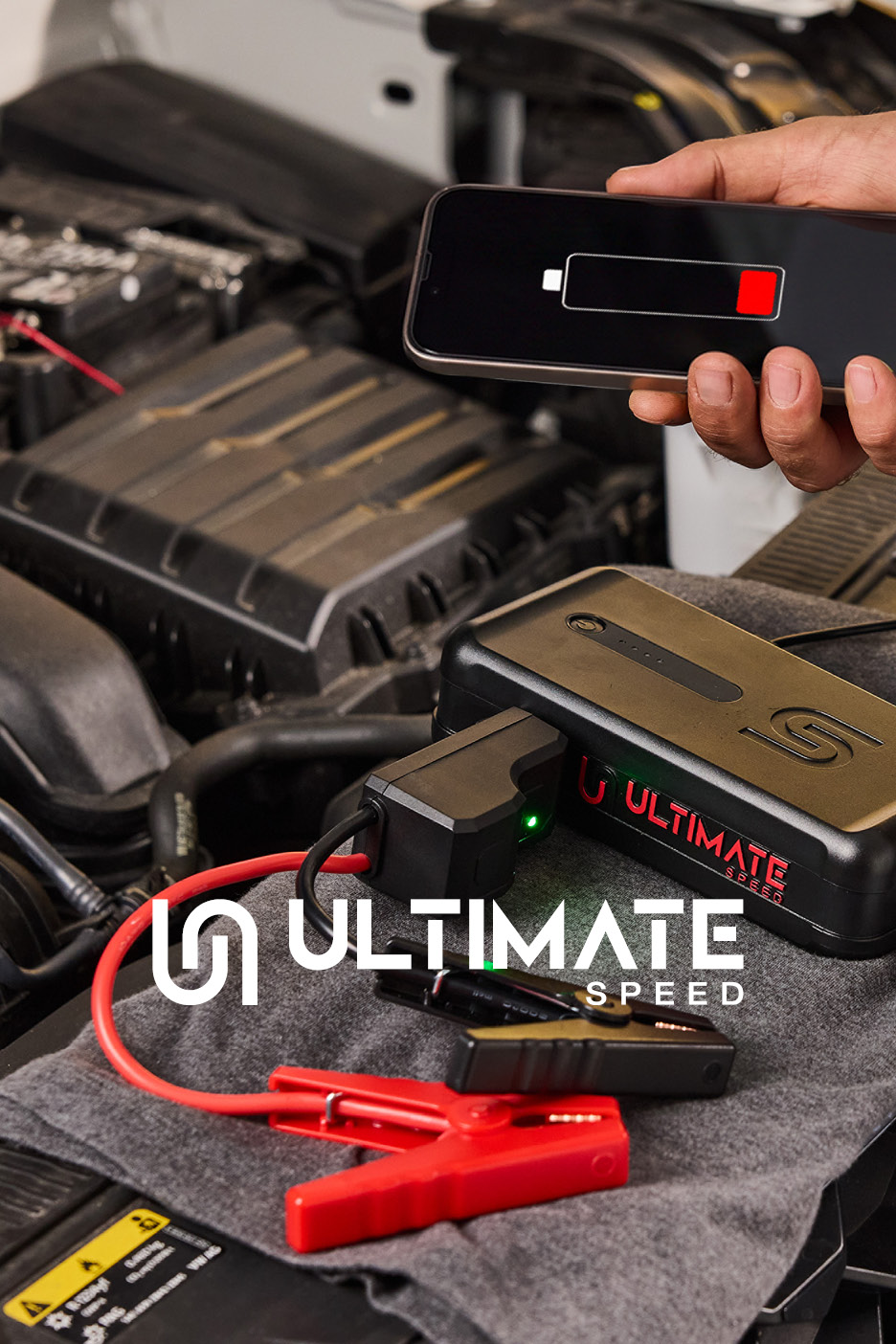 Avviatore d'emergenza per auto con powerbank Ultimate Speed posizionato nel vano motore, con pinze di collegamento e uno smartphone con batteria scarica in mano.